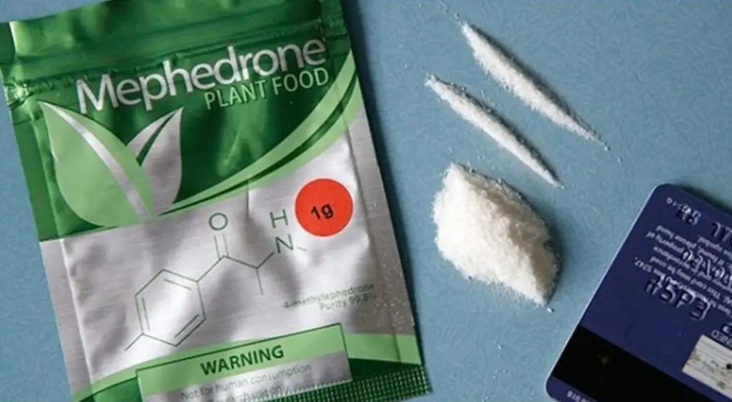 Green Mephedrone Plant Food package with a chemical diagram and a red '1g' label, next to white powder lines on a blue surface, plus a blue card nearby.