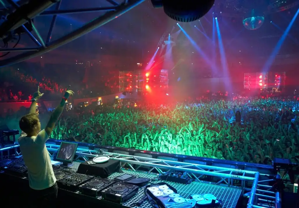 DJ on stage manipulating mixer while a large crowd cheers with colorful lights and a disco ball overhead